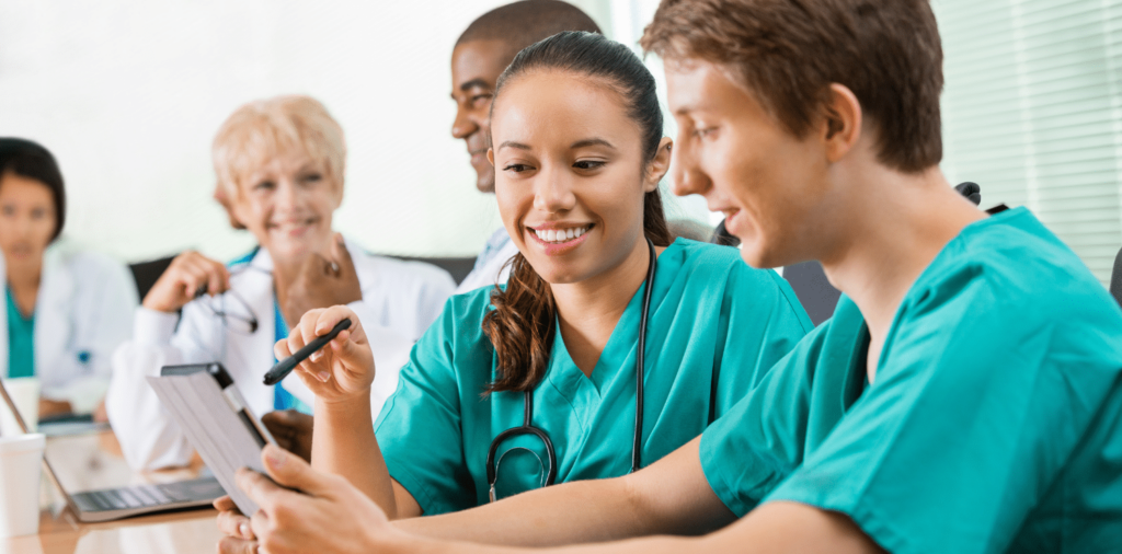 Two happy nurses in scrubs discussing their first travel nurse assignment while looking at a tablet, with other medical professionals in the background smiling and engaged in conversation.