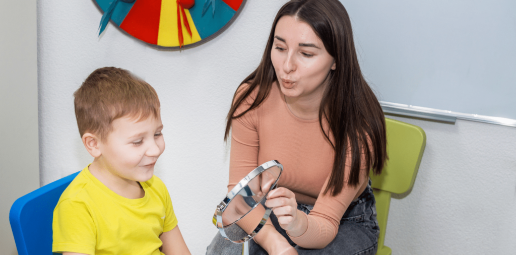 A Speech Language Pathologist (SLP) sits next to a young child. The SLP is holding a mirror. The child, with a look of curiosity and excitement, is attentively listening to the SLP's words. The SLP uses various tools and gestures to engage the child in communication exercises, encouraging them to mimic words. Through their interaction, the SLP created a supportive and nurturing environment to help the child develop their speech and language skills. Speech pathology