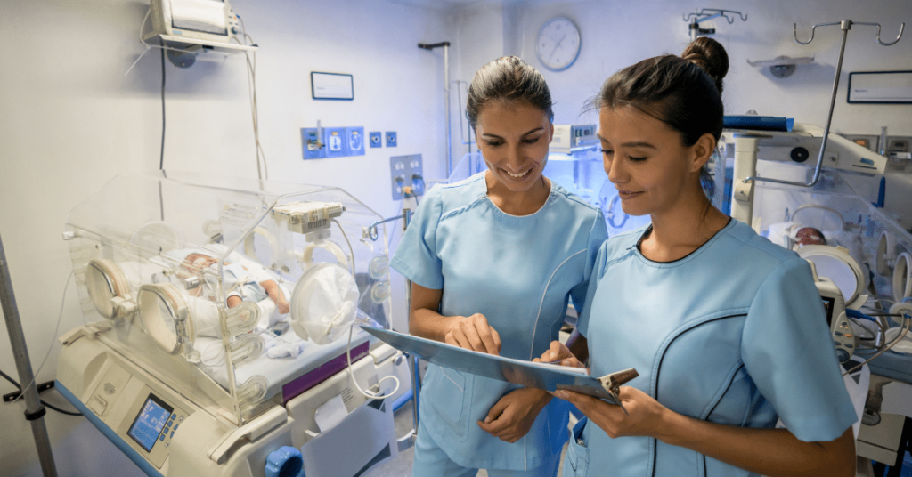 Neonatal nurses in the NICU smile while reviewing a premature baby's medical chart. Incubators with tiny infants fill the background.