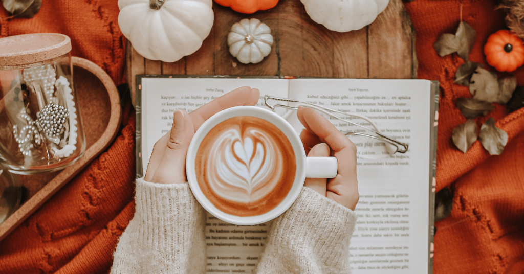 A woman sits at a cozy table adorned with pumpkins, holding a warm autumnal coffee in one hand and a book in the other. She is engrossed in her reading, creating a warm and inviting atmosphere. This image embodies ways to make your temporary home feel cozy during Thanksgiving, especially for travel nurses and allied health professionals residing in temporary accommodations during the holiday season.