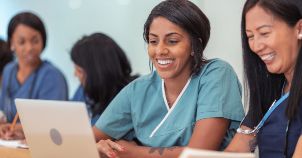 Female nursing students attending class together. They are seated at a long table in a classroom, collaborating in pairs on a project. Furthering education provides career advancement for nurses.