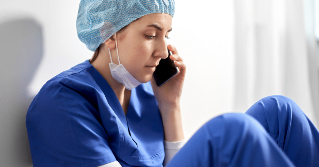 A young nurse appears distressed, sitting on the floor with her back against a wall. She's holding a phone to her ear, seemingly in the midst of a serious conversation. Her expression and posture convey a sense of sadness and frustration, suggesting she's talking to her recruiter about her dissatisfaction with her current travel nursing assignment. The environment around her is nondescript, putting the focus on her emotional state.