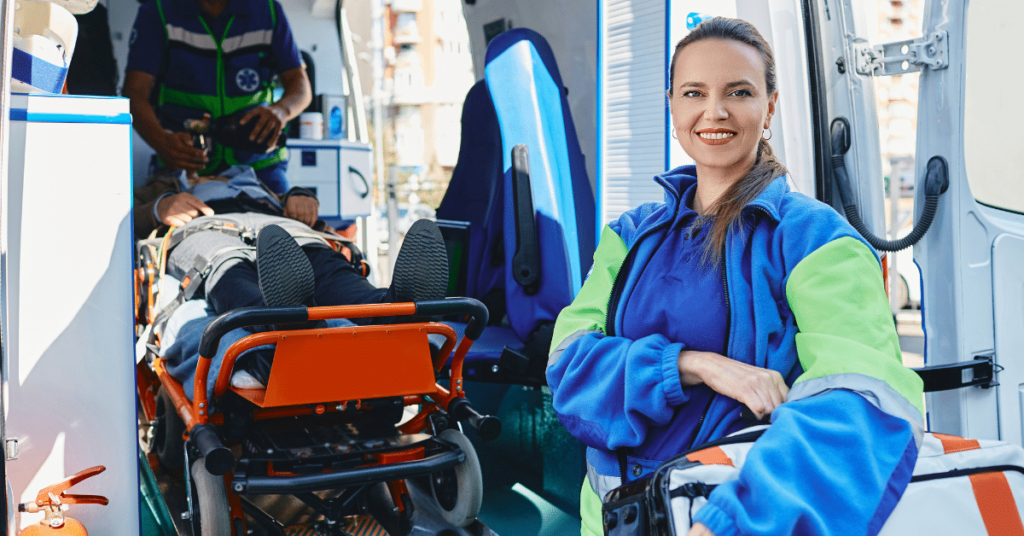 An emergency medical technician, with a warm and reassuring smile, stands outside of an ambulance. In the background, her partner, also an EMT, is inside the ambulance, diligently attending to a patient, providing essential medical care.