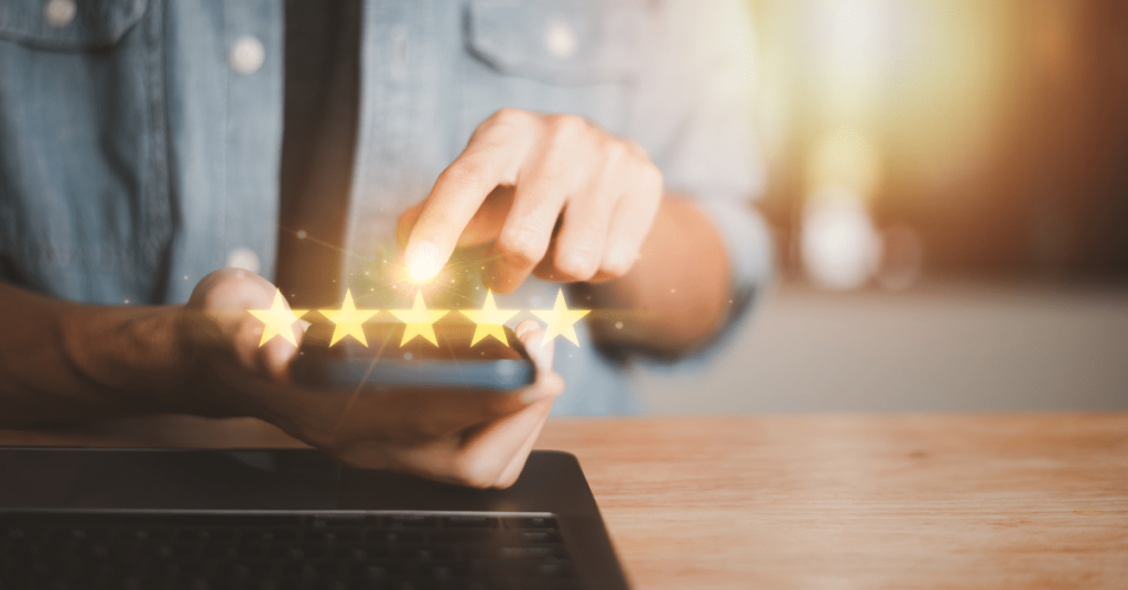 Close-up image of a man's hand holding a cellphone, with the screen clearly displaying a 5-star review being selected. The man's pointer finger is shown pressing one of the five brightly lit star icons on the screen, indicating a positive rating. The background is blurred, focusing the attention on the action of giving a 5-star review.